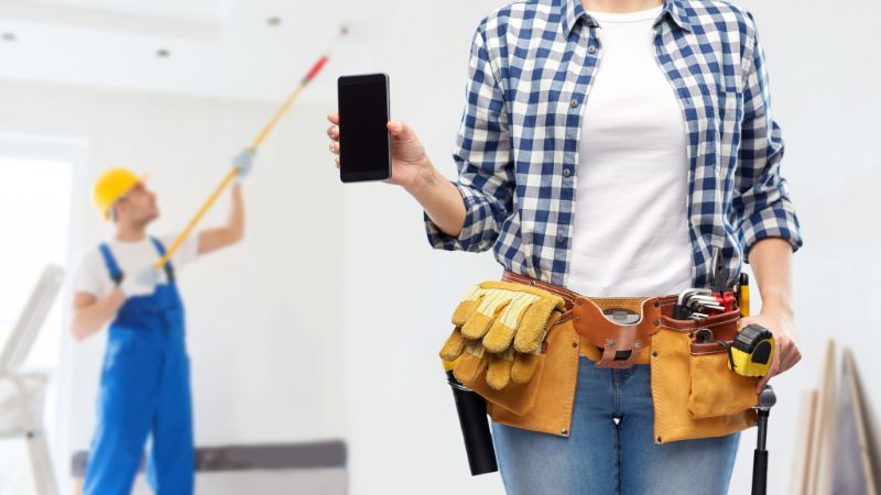 femme avec smartphone et ceinture outils de travail avec dans le fond un peintre peignant un plafond